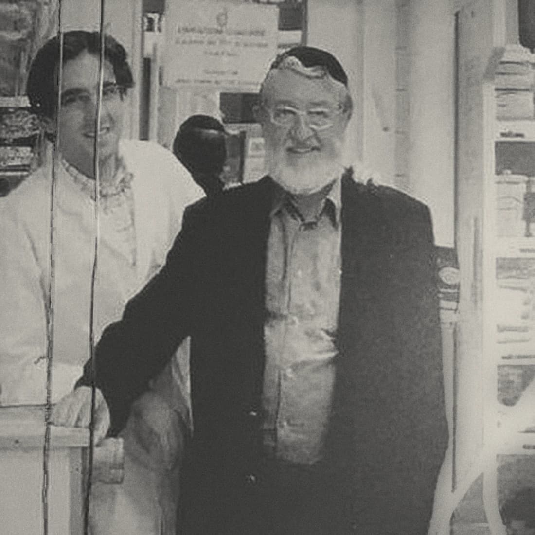 Black and white archive photograph: two men behind the counter of a delicatessen, one in a white coat, the other in a suit, in front of the shop shelves